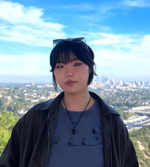 Young person with bangs and sunglasses on head, neutral expression, city skyline and blue sky