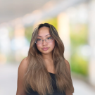 Young woman with long light-brown hair and glasses, neutral expression, blurred outdoor background